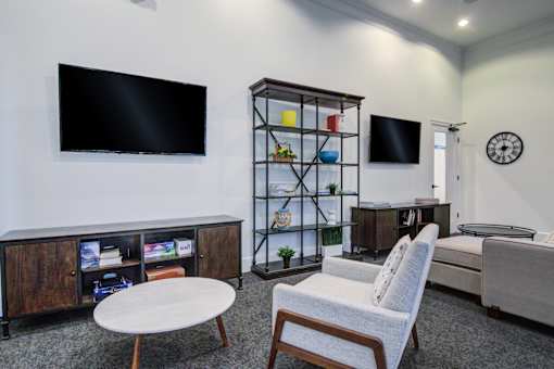 A living room with a white chair, a white table, a wooden cabinet, a glass cabinet, a flat screen TV, and a clock on the wall at Sovereign at Overland Park Apartments, Overland Park