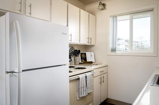 A white refrigerator stands in a kitchen with white cabinets and a window.