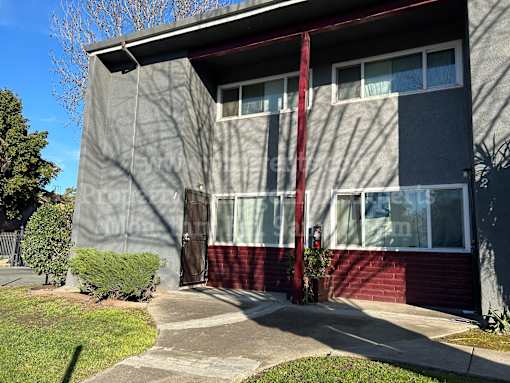 A grey and red building with a red awning and a small tree in front.
