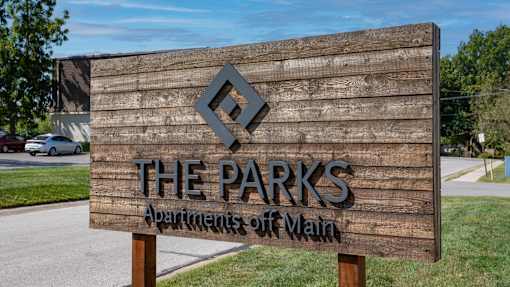 A wooden sign for "The Parks Apartments off Vlaan" stands in front of a parking lot.