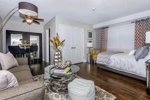 A living room with a grey couch, a glass coffee table, and a bed with a white comforter.