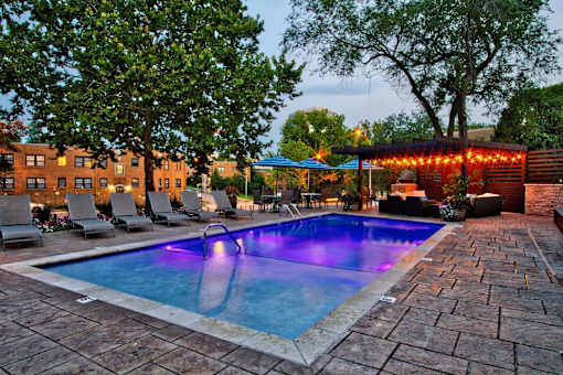 A pool surrounded by chairs and trees with lights on the building.