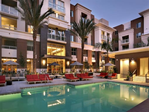 A large swimming pool is surrounded by red chairs and palm trees in front of a building. at West Park Apartments, San Diego, CA, 92108
