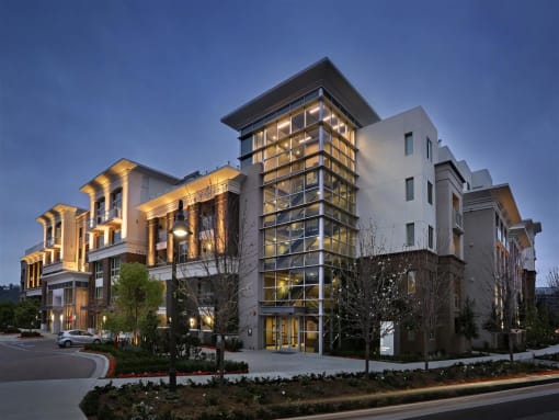 A modern multi-story building with a mix of glass and solid exterior walls is illuminated from within and surrounded by a landscaped area at West Park Apartments, San Diego, CA, 92108
