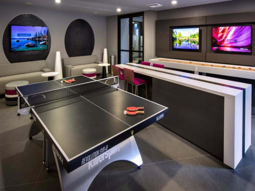 A ping pong table is in the middle of a room with a bar and two TVs on the wall at West Park Apartments, California
