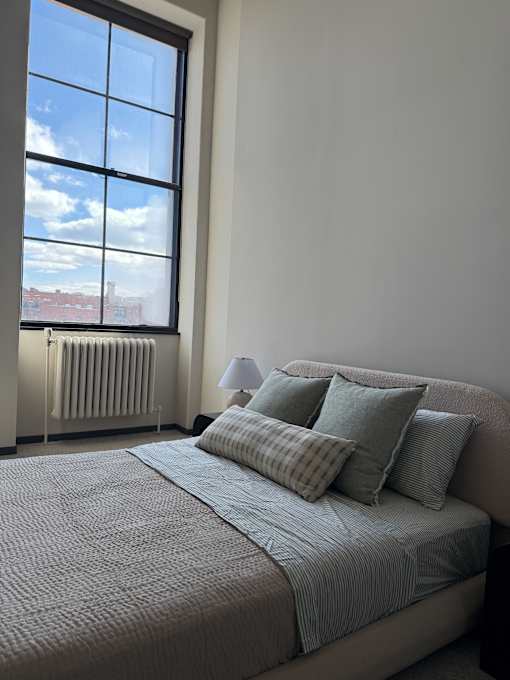 A bed with a grey comforter and pillows is in front of a window.