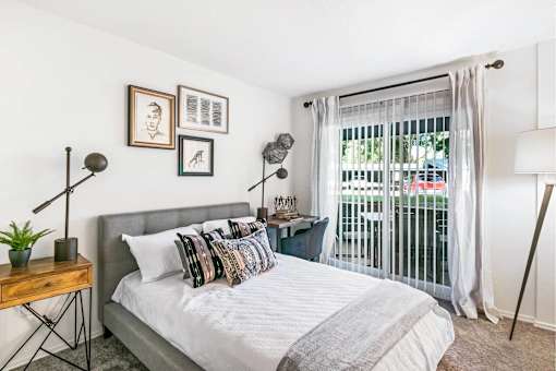 A bedroom with a bed, a nightstand, and a window with a view of a balcony.