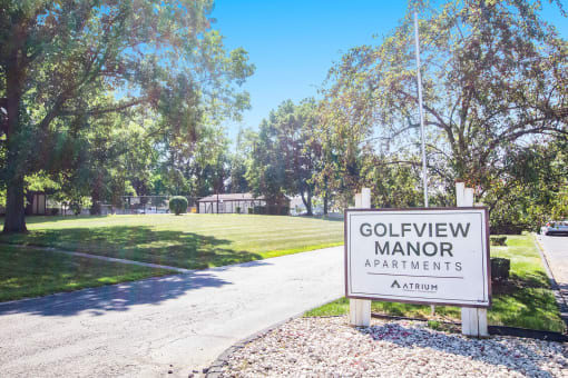 A sign for Golfview Manor Apartments stands in front of a tree-lined street.