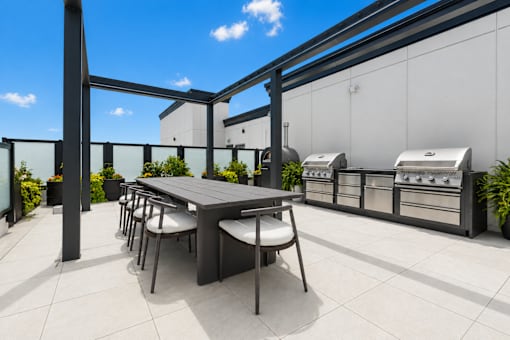 A table and chairs are on a patio with a grill and a building in the background.