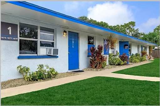 A white building with blue doors and windows has a sign on the left side.