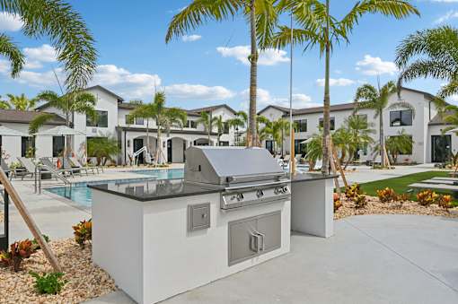 A modern outdoor kitchen with a grill and sink is set up in front of a row of houses at The Hadley - North Port, FL Apartments, North Port