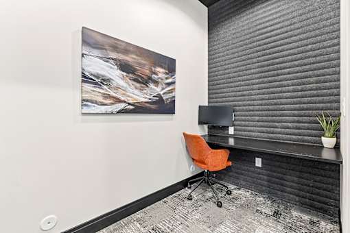 A modern office with a large painting on the wall and a black desk with an orange chair.