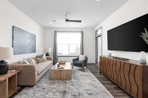 A modern living room with a beige sofa, a grey rug, a wooden coffee table, a black and white painting, a grey armchair, a wooden sideboard, and a large window.