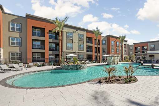 A swimming pool surrounded by palm trees and lounge chairs in front of apartment buildings.