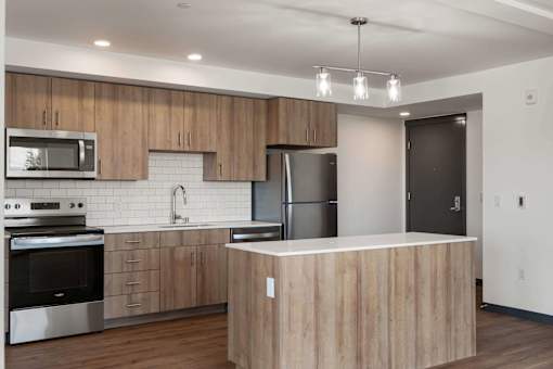 White Center, WA Studios For Rent - Elevation16 - Kitchen With Stainless Steel Appliances, Vinyl Plank Flooring, Kitchen Island, and Generous Kitchen Storage With Hardwood Cabinets