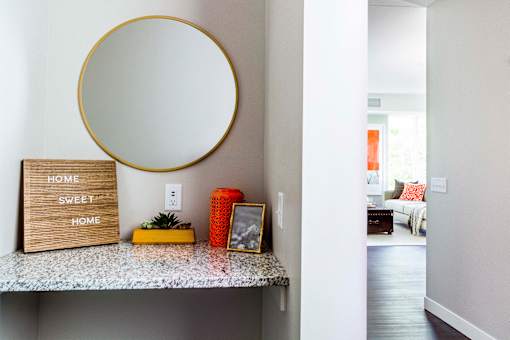 Entrance to a modern apartment: A sleek hallway vanity with a granite countertop welcomes guests, seamlessly transitioning into the well-lit living room flooded with natural light.