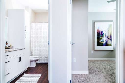 Modern apartment bathroom featuring stain-resistant floors and elegant granite countertops. The adjacent spare bedroom boasts plush carpeting and serene nature views.