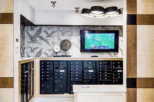Interior view of a modern apartment complex mail room featuring rows of individual mailboxes neatly arranged along the wall. In the center, there is a flat-screen TV displaying important community announcements and updates.