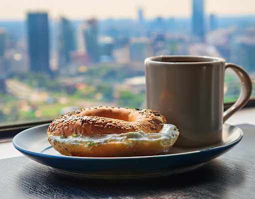 A bagel with cream cheese and a coffee mug on a table with a city view in the background.