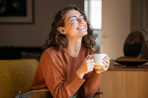 A woman with curly hair is wearing glasses and a brown sweater, smiling while holding a white mug.