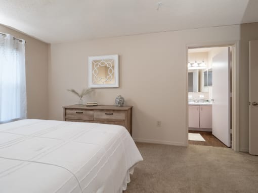 a bedroom with a white bed and a bathroom with a sink at The Fountains Apartments, Pensacola, FL