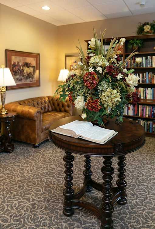 A room with a brown couch, a round table with a book on it, and a flower arrangement.