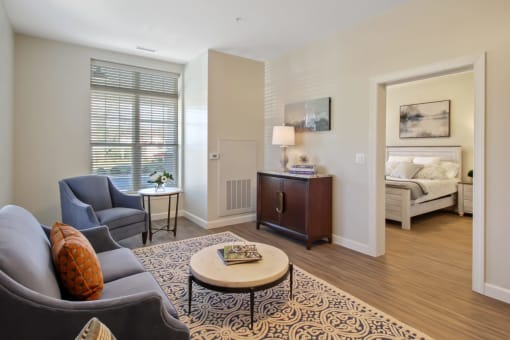 A living room with a grey couch, a coffee table, and a bed in the background at White Springs Senior Living, Virginia 20186