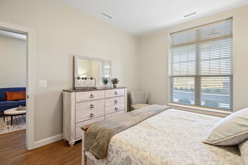 A bedroom with a large bed, a dresser, and a window with blinds at White Springs Senior Living, Warrenton, Virginia