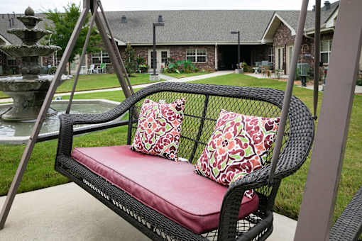 A black wicker swing with red cushion and pillows is in the foreground with a house and fountain in the background.