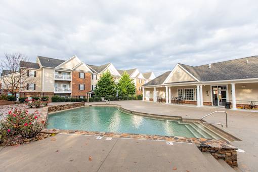 A swimming pool in front of a building with a patio and chairs.