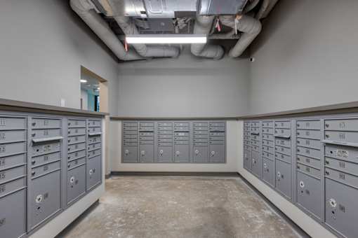A room with a concrete floor and grey walls has a number of grey lockers with numbers on them.