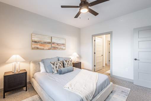 A bedroom with a bed, two lamps, a ceiling fan, and a painting on the wall.