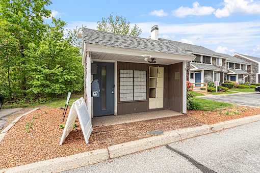 A small house with a grey roof and a brown door is surrounded by a gravel area and a grassy area.