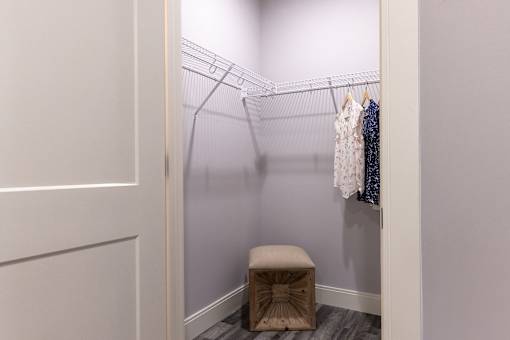 A small closet with clothes hanging on a wire rack and a stool in front.