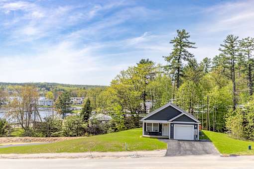A house with a garage is situated on a grassy area with trees and a body of water in the background.