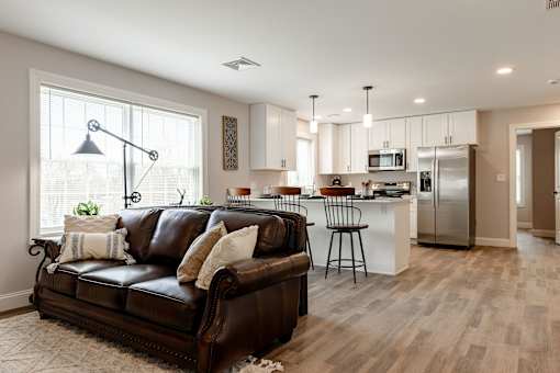A brown leather couch with white pillows sits in a living room with a kitchen in the background.