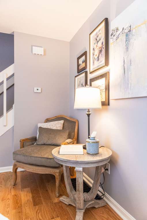 A table with a lamp and a book on it with a chair and a painting in the background.