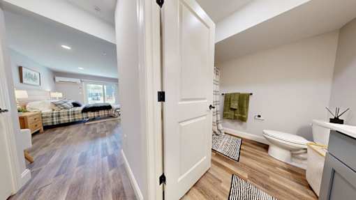 A bathroom with a toilet, sink, and a bedroom with a bed and dresser.