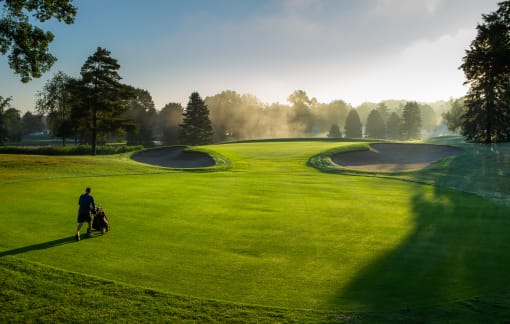a golfer walks across a green with a bunker in front of him