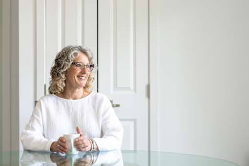 A woman with glasses and curly hair is sitting at a table with a white mug in her hands.