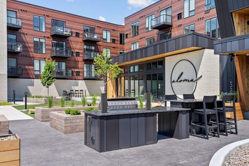 A modern outdoor seating area with a bar and chairs is situated in front of a building with a sign that reads "Alma.".