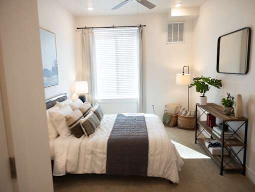 A bedroom with a bed, a window with blinds, a ceiling fan, a mirror, and a shelf with a plant and other items.