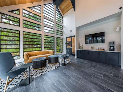 A community lounge with a sofa, two chairs, coffee tables, and large windows near a resident coffee station with a TV.