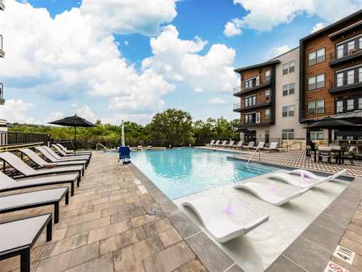 A swimming pool with lounge chairs and umbrellas near large trees and apartment buildings.