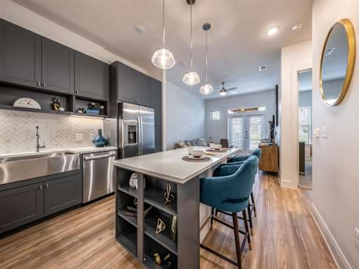 A kitchen with an island, a large farmhouse-style sink, stainless steel appliances, and pendant lights near a living room and bedroom.