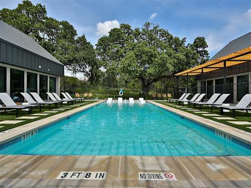 A gated swimming pool lined with lounge chairs near a clubhouse and large trees.