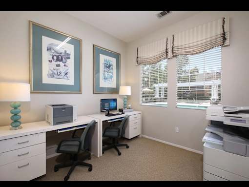 A well-lit office with two desks and chairs, a printer, and two framed pictures on the wall.