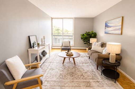 A living room with a grey rug, a grey sofa, a wooden chair, a wooden coffee table, a white side table, a white wall, a window, a plant, and a painting.