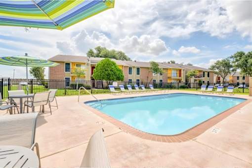 A pool surrounded by chairs and umbrellas in front of apartment buildings.