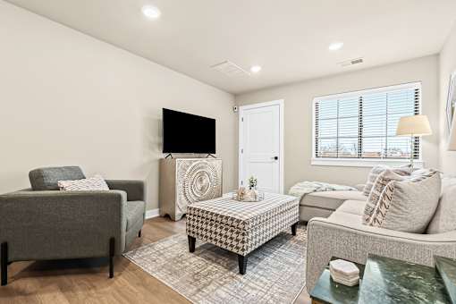 A living room with a grey couch, a black and white ottoman, and a flat screen TV.